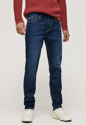 Mężczyzna w ciemnoniebieskich dopasowanych dżinsach, białych sportowych butach i czerwonym swetrze z dzianiny, z rękami swobodnie opuszczonymi wzdłuż ciała.