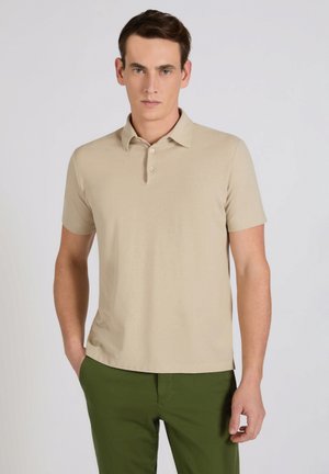 Man draagt een beige poloshirt met korte mouwen en groene broek, staat met één hand in zijn zak tegen een effen lichte achtergrond.