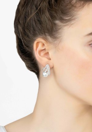 Pendientes de tono plateado que presentan un diseño fluido con acentos en forma de ola y una gran perla blanca en la base. Textura suave con detalles brillantes.