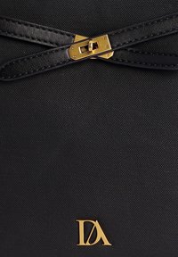 Material texturizado negro con un cierre dorado y correas de cuero negro. Presenta un logo dorado en la esquina inferior derecha.