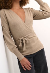 Beige wrap sweater in soft knit fabric, featuring a deep V-neck, long puff sleeves, and a tie belt at the waist. Paired with dark jeans.