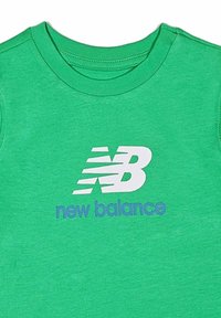 T-shirt verde in cotone con scollatura rotonda, caratterizzato da un logo "new balance" bianco e blu stampato sulla parte anteriore. Texture morbida, design casual.