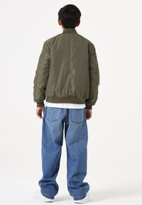 Olivgrüne Bomberjacke mit geripptem Kragen und Bündchen, die eine gepolsterte Textur aufweist. Getragen mit hellblauen, locker sitzenden Denim-Jeans.