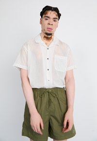 Camisa de botão de manga curta, branca com riscas, com um bolso, combinada com calções verde-oliva com uma cintura elástica e cordão.