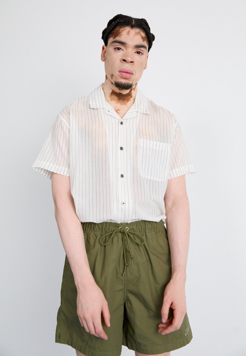 Kurzärmliges, gestreiftes, weißes Hemd mit Knopf und einer Tasche, kombiniert mit olivgrünen Shorts mit elastischem Bund und Kordelzug.