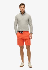 Pullover grigio a maniche lunghe con colletto a mezza zip, abbinato a pantaloni corti arancioni vivaci e sneaker beige, creando un outfit casual.