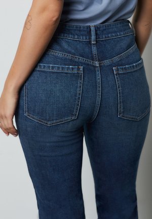 Osoba nosi tamnoplave traperice visokog struka s stražnjim džepovima, vidljiva ruka s cvjetnim tetovažom i svijetloplavu košulju uvučenu unutra.