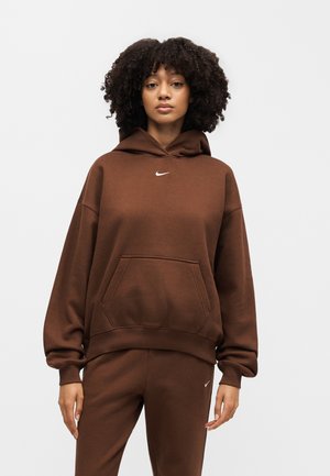 Kvinde med krøllet hår iført en brun Nike hættetrøje og matchende joggingbukser, stående mod en ensfarvet hvid baggrund.