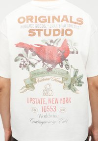 Πίσω μέρος λευκού πουκαμίσου με κόκκινο καρδινάλιο σε κλαδί, κείμενο «Originals Studio», «Hudson Valley Natural Scene» και «Upstate, New York 10553».