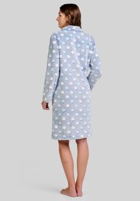 Mujer de pie descalza, de espaldas, vistiendo una bata de felpa ligera de color azul claro hasta la rodilla con lunares blancos y azules sobre un fondo gris.