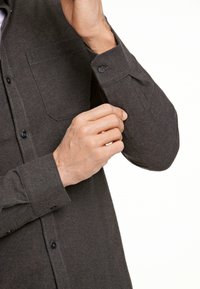 Camicia grigia scuro con tessuto strutturato, dotata di taschino sul petto, maniche lunghe arrotolate ai polsi e cuciture a contrasto.