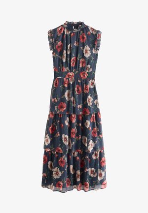 Vrouw loopt binnenshuis, draagt een mouweloze marineblauwe midi-jurk met bloemen, een geplooide rok en beige hoge hakken sandalen.