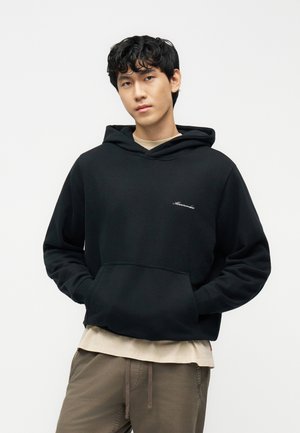 Νεαρός με σγουρά μαλλιά που φοράει μαύρο φούτερ με κουκούλα πάνω από μπεζ πουκάμισο και καφέ παντελόνι, όρθιος με τα χέρια στις τσέπες του φούτερ.