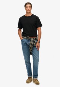 Sort kortærmet T-shirt, blå jeans, ternet skjorte bundet om taljen, lysebrune ruskindsko og et armbåndsur, stående på en ensfarvet baggrund.