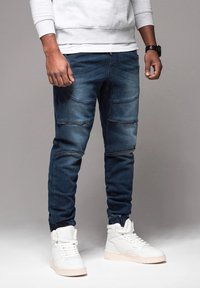 Joggers in denim blu scuro con dettagli a coste, chiusure con zip e polsini elastici, abbinati a sneakers bianche alte.
