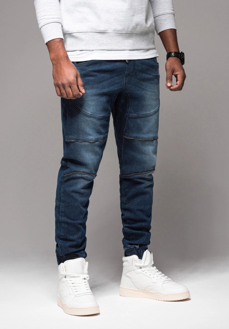 Joggers in denim blu scuro con dettagli a coste, chiusure con zip e polsini elastici, abbinati a sneakers bianche alte.