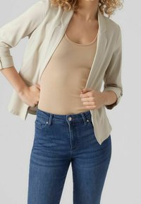 Femme portant un blazer beige sur un débardeur côtelé couleur sable et un jean bleu taille haute, ajustant le blazer avec les mains à la taille.