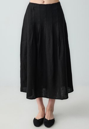 Jupe en lin noire avec des plis, un design long et fluide, et un ourlet qui se situe légèrement au-dessus des chevilles. Associée à des chaussures plates noires.