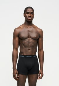 Μυώδης άνδρας μοντέλο με σκούρα επιδερμίδα, όρθιος, φοράει μαύρα εσώρουχα Calvin Klein boxer briefs, σε απλό φωτεινό φόντο.