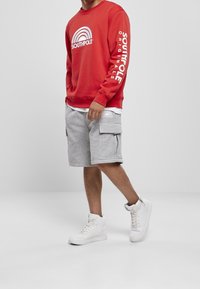 Uomo che indossa un felpa a maniche lunghe rossa con il logo "Southpole", pantaloni cargo grigi con logo e sneaker alte bianche, in piedi su un pavimento grigio.