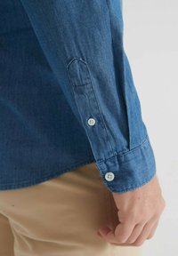 Manche de chemise en denim bleu avec des boutons blancs au poignet, portée par une personne à la peau claire et portant un pantalon beige.