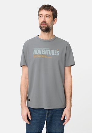 Mann mit Bart, der ein graues T-Shirt mit dem Schriftzug "ADVENTURES" und blaue Jeans trägt, steht vor einem einfarbigen weißen Hintergrund.