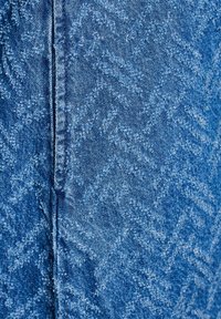 Jeanstyg i ljusblått med ett strukturerat zickzackmönster. Detalj av sömnadsstygnen längs sömmen är synlig.