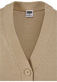 Cardigan beige en tricot avec col en V côtelé, fermeture par un seul bouton, et une étiquette "Urban Classics" indiquant XS, fabriqué au Bangladesh.