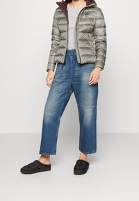 Veste grise matelassée avec une doublure bordeau, associée à un jean bleu large en denim et des chaussures noires sans lacets. Comprend une fermeture éclair et des poches.