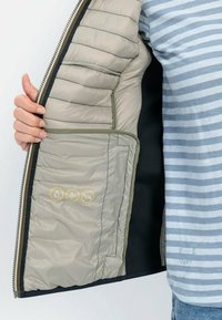 Lichtgewicht beige jack met gequiltte textuur, voorzien van een zwarte voering en binnenzak. Symbolen duiden op waterafstotende en dons vullingseigenschappen.
