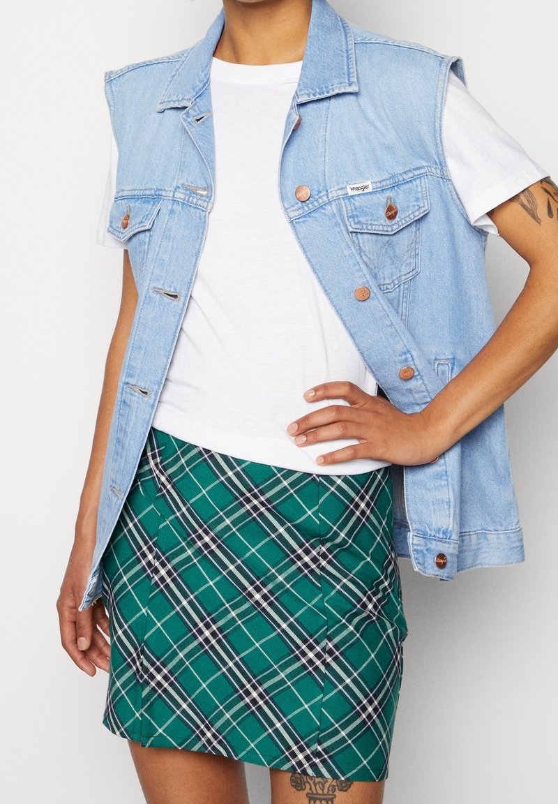 Gilet en denim bleu clair avec des boutons en cuivre, t-shirt blanc et mini jupe écossaise verte avec des accents noirs et blancs.