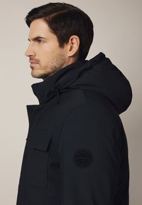 Czarna kurtka parka z kapturem, z zapięciem na zamek, przednią kieszenią z klapką oraz okrągłą naszywką z logo na rękawie, gładka tekstura i dopasowany krój.