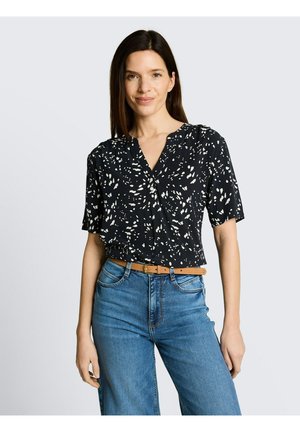 Vrouw met lang bruin haar, die een zwart kort mouwblouse met patroon, blauwe high-waisted jeans en een beige riem draagt, staand tegen een effen achtergrond.