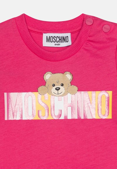 La camiseta rosa de manga corta para bebé presenta un gráfico de un oso sobre el logo "MOSCHINO", con dos botones a presión en el hombro. Material de algodón suave.