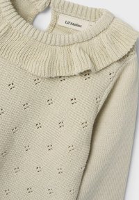 Hellbeige gestrickter Pullover mit Rüschensaum, der ein strukturiertes Muster aus kleinen Punkten und einen offenen Rücken aufweist. Das Etikett trägt die Aufschrift "Lil'Atelier."