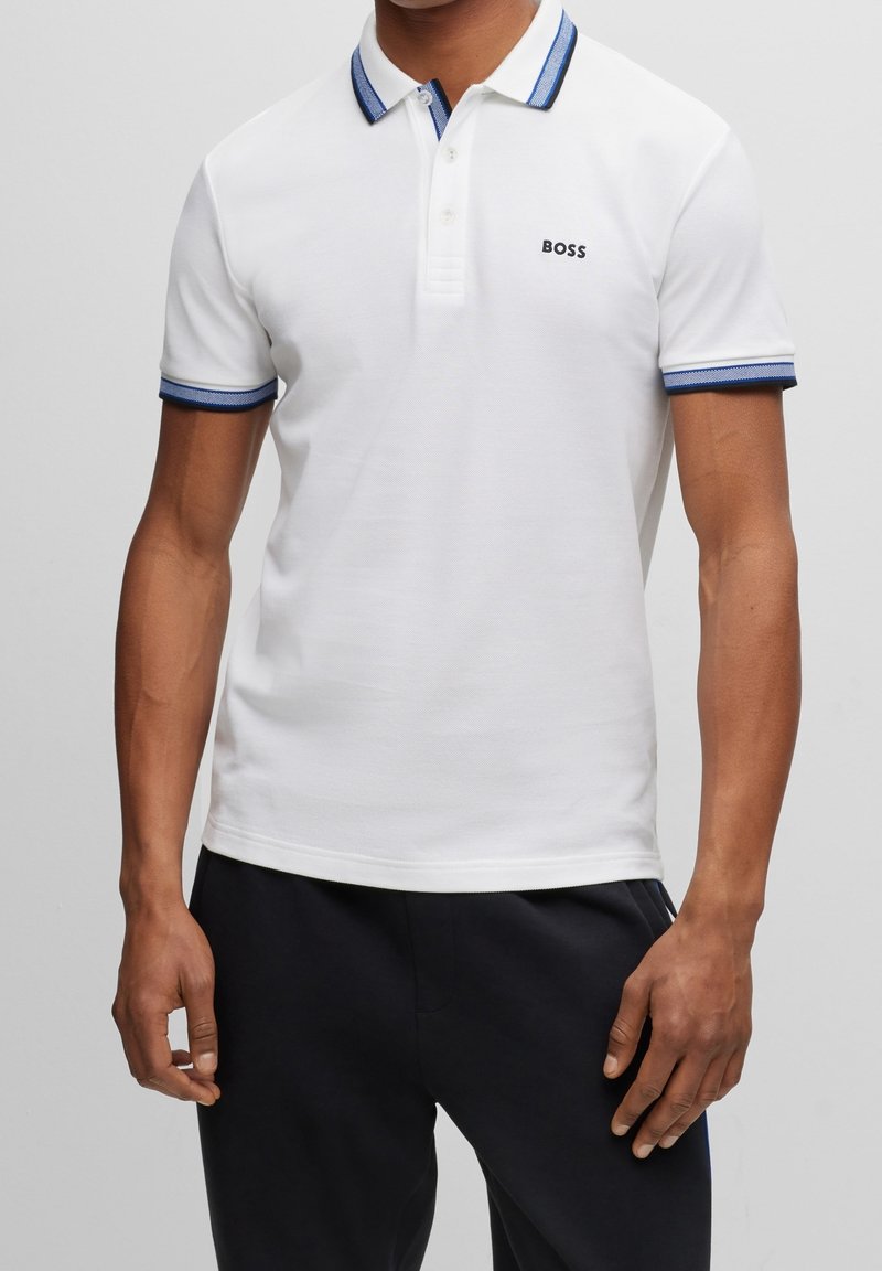 Homme portant un polo blanc avec col et bordures de manches bleu, logo "BOSS" sur la poitrine gauche, associé à un pantalon noir.