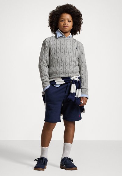 Grauer Strickpullover, blaues Hemd mit Kragen, marineblaue Shorts, Kniesocken und marineblaue Schuhe. Ein gestreifter Pullover um die Taille gebunden.