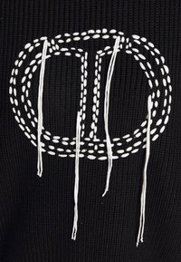 Tissu noir texturé présentant un logo brodé blanc avec des fils blancs lâches et allongés accentuant le motif.