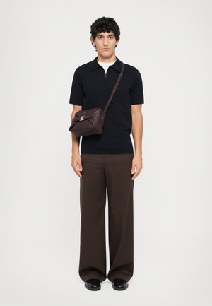Bolso cruzado marrón con cierre de solapa, herrajes plateados y cuero texturizado. El modelo lleva una polo negra, pantalones anchos marrones y zapatos negros.