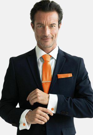 Dunkelblauer Anzug mit weißem Hemd, orangefarbener Krawatte und Tasche täschchen. Weißer Manschettenknopf mit orangem Detail, silberner Krawattenclip, glatte Stofftextur.