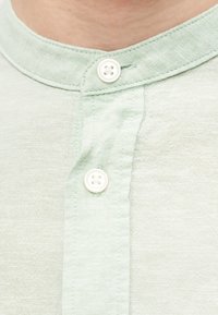 Κοντινό πλάνο στο λαιμό ενός ανοιχτοπράσινου πουκάμισου henley με δύο λευκά κουμπιά δεμένα στο μπροστινό άνοιγμα.