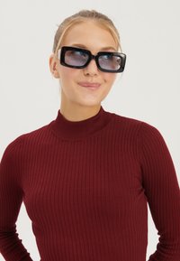 Černé oversized sluneční brýle s gradientovými skly, které doplňují žebrovaný burgundský top s krátkým stojáčkem. Tkanina vypadá lehce a těsně přiléhající.