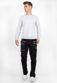 Felpa grigia, pantaloni cargo neri con molte tasche con cerniera e dettagli in cintura, scarpe bianche, in piedi contro uno sfondo bianco.