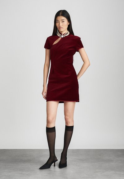 Rochie din catifea bordeaux cu mâneci scurte și un design cu decupaje. Asortată cu ciorapi negri semi-transparenti până la genunchi și pantofi cu toc ascuțit negri.