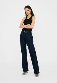 Tumed sinised denim teksad kõrge vöökoha, sirge sääre lõike ja kontrastsete õmblustega. Kanda musta lühikeste varrukatega topi ja mustriliste kontsakingadega.