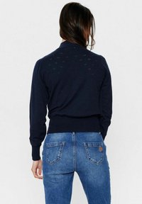 Navyblauwe trui met gedetailleerde gaatjessteek aan de achterkant, geribbelde manchetten en zoom. Gecombineerd met blauwe spijkerbroek met achterzakken.