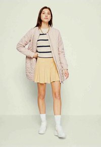 Giovane donna che indossa una giacca trapuntata rosa, una maglietta a righe, una gonna plissettata beige, calze bianche e sneaker bianche su uno sfondo neutro.