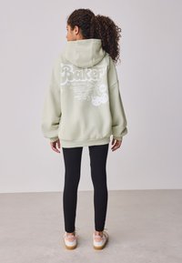 Túlméretezett világoszöld kapucnis pulóver fehér grafikus szöveggel és díszítésekkel, fekete leggingsszel és fehér, rózsaszín részletekkel ellátott sportcipővel párosítva.