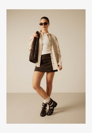 Naine, kes kannab päikeseprille, üle suurusega triibulist särk, valget topi, musta miniseelikut, sandaale sokkidega, suuri rõngaskujulisi kõrvarõngaid, hoides tumedat kotti.