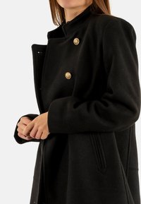 Abrigo negro de mezcla de lana con diseño de doble botonadura, botones dorados, cuello alto y acentos de costura vertical en los lados.